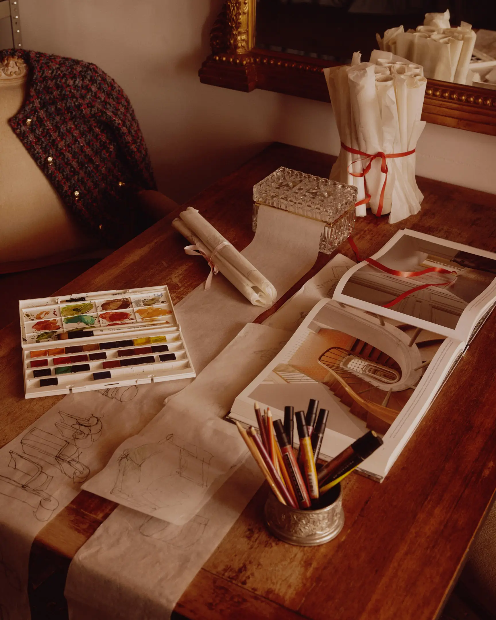 A table with drawing materials, Hermes purse, and jacket with an ornate mirror, and studio shelving in the background. The studio of Lulu LaFortune, a designer based in Los Angeles, Ca. Shot originally for House Beautiful publication. 